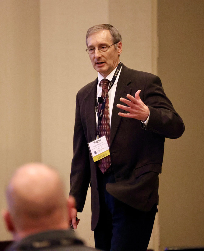 A man in a suit speaks passionately at a conference, gesturing with one hand. A lanyard and badge are visible. Audience member in foreground.