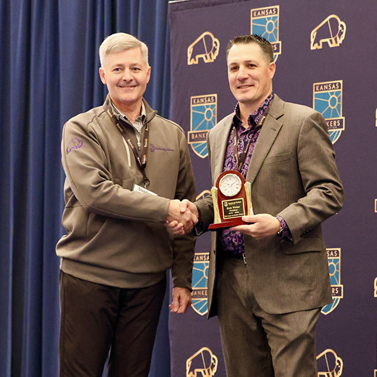 2026 Kansas Ag Bankers Conference; Two men stand on stage shaking hands; one holds a plaque with a clock. They're in business attire with a backdrop of Kansas Bankers logos.