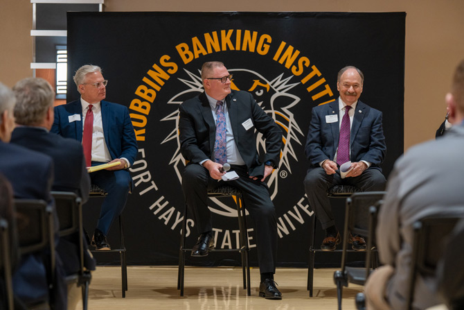 Three men in suits sit on chairs in front of a Robbins Banking Institute banner. They appear engaged in a panel discussion, with an audience seated in front.