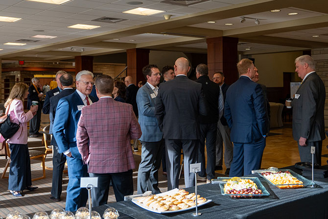 A group of people in business attire engage in conversation at a networking event. Tables hold snacks like cookies and fruit under bright ceiling lights.