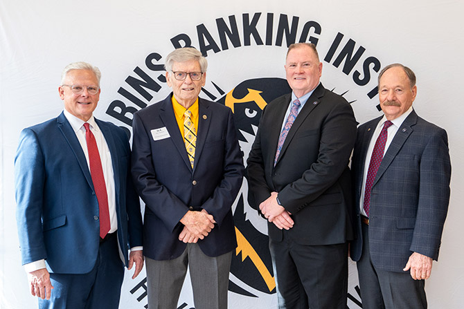 Four men in suites smile in front of a wall with the Robbins Banking Institute logo