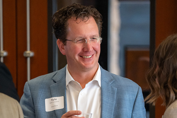 Man in a blue suit jacket smiles while holding a glass, standing in an indoor setting. He wears a name tag and appears to be engaged in conversation.