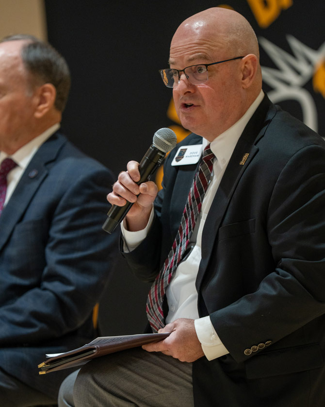 A man in a suit and tie is seated, holding a microphone, engaged in speaking. He has a focused expression. Another man is partially visible beside him.