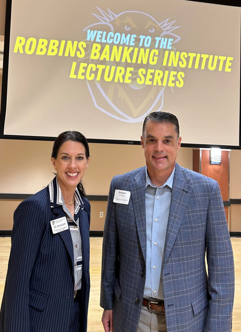 Two people smiling in business attire in front of a screen displaying "Welcome to the Robbins Banking Institute Lecture Series." Professional setting.