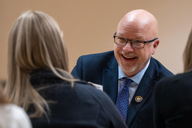 A smiling bald man in a suit and glasses converses with two women, creating a friendly atmosphere. He wears a name tag and a pin, indicating professionalism.