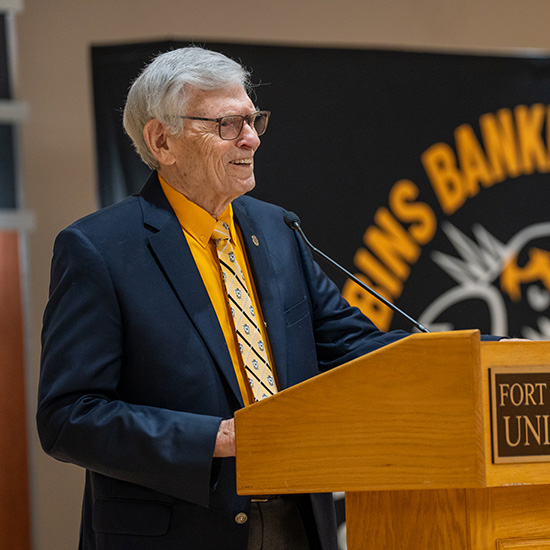 2026 Robbins Banking Institute Lecture Series Highlights; Elderly man with white hair and glasses speaks at a podium. He's wearing a navy blazer and yellow shirt. A banner behind reads "FORT...UNIV."