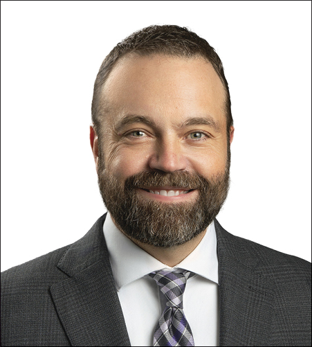 Ben Drouhard; A man with a beard and short hair smiles warmly in a professional headshot. He wears a dark suit, white shirt, and patterned tie against a white background.