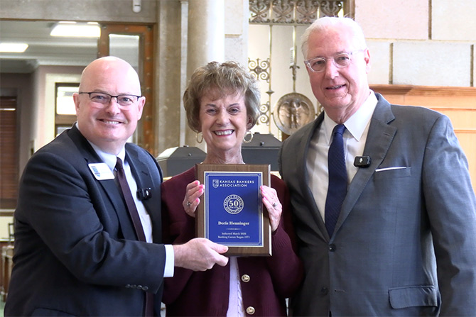 Three people stand together smiling indoors. The woman in the center holds a blue and gold plaque. The mood is celebratory and professional.